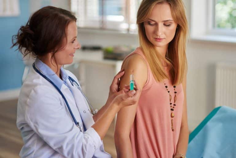 Allergy Treatment Immunotherapy Woman in a doctors office getting a shot to the arm from her doctor. She is looking down at her arm as her doctor looks at the her with her thumb over the plunger.