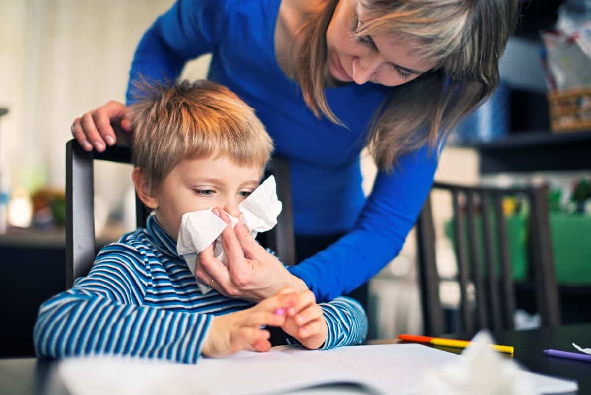 Pediatric-Allergy-symptoms Mother leaning over child in kitchen chair as they hold a tissue up to a sitting child's nose
