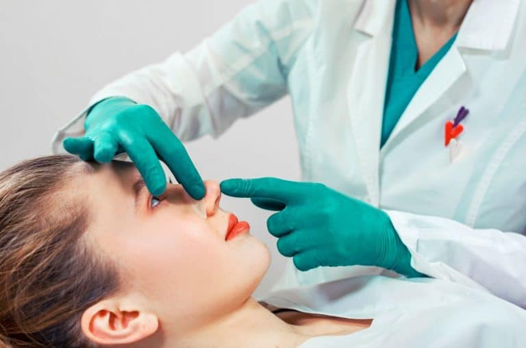 Facial-Plastics-Rhinoplasty-Consultation Image starts at just below chest height. Woman is laying down in medical outfit. The upper body and arms of a doctor wearing scrubs is connected to gloved hands which are placing a piece of medical tape onto the womans nose as she looks up at them from the table.